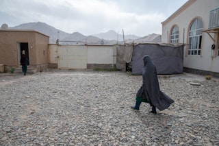 A veiled woman walks on stones towards a small building.