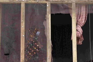 A masked and veiled young woman in coloured outfit stands, barely visible through a broken screened window.