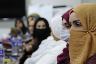 A woman in a mustard headscarf looks left in the foreground, with a room full of blurred women in the background.