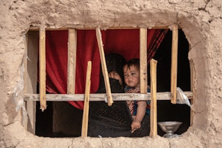 A woman in a black hijab holds her child near a red curtain looking out a boarded-up window in a clay home.