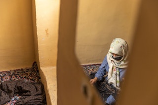 A veiled young woman crouches on the floor, as seen through the reflection of a mirror on the wall.