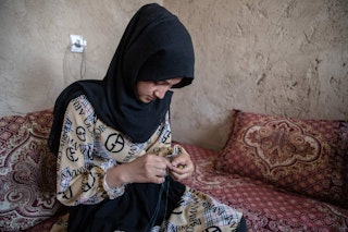A veiled young girl sits sewing on a daybed.