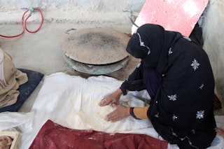 A woman kneels to press flour onto a bedsheet on the floor, preparing bread.