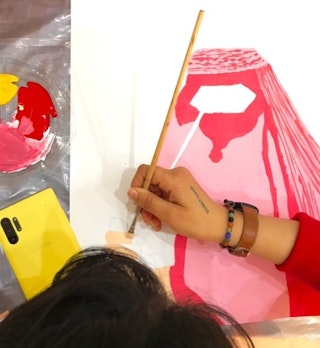 A woman paints a woman in a pink and red burqa.