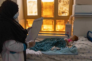 A female health professional looks at the chart of a female baby, which lies in the hospital bed next to her.