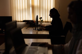 Two women with headphones on, sit at their desks and computers, working in dim light.