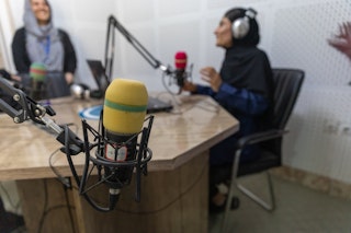 A yellow microphone in foreground, a woman smiles while sitting on air in a radio station in the background.