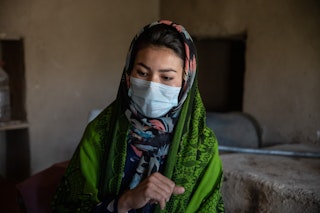 A young woman in a facemask and green veil sits in her home.