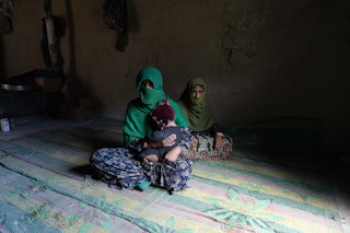 A woman in a green veil sits, a toddler on her lap and an older daughter in the background.