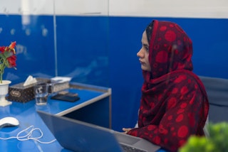 A young woman in a red spotted hijab sits behind a blue desk.