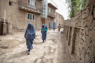 Two women in blue burqas walk down a narrow alley, one holding a small child.