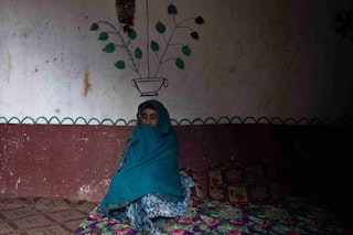 A veiled older woman sits next to a wall with a flower painted on it.