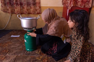 A mother sets a pot of water to boil over a gas cylinder, her young daughter visible next to her, from a side angle.