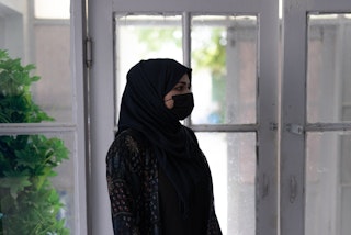A woman in a black facemask and hijab stands by french doors, looking to the right.