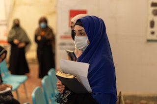 Veiled young woman holds books in a meeting space, wearing a facemask.