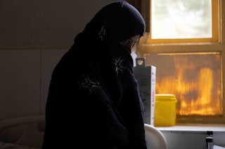A woman standing near a window peaks out from behind a black hijab.