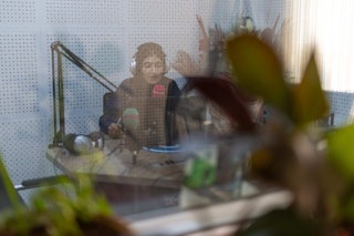 A woman on the air at a radio station visible behind blurry flowers in the foreground.