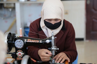 A veiled and masked woman sits threading a sewing machine.