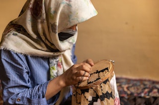 A veiled and masked girl sits embroidering.