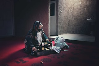 A veiled and masked girl sits at a sewing machine on a red carpet in a dimly lit room.