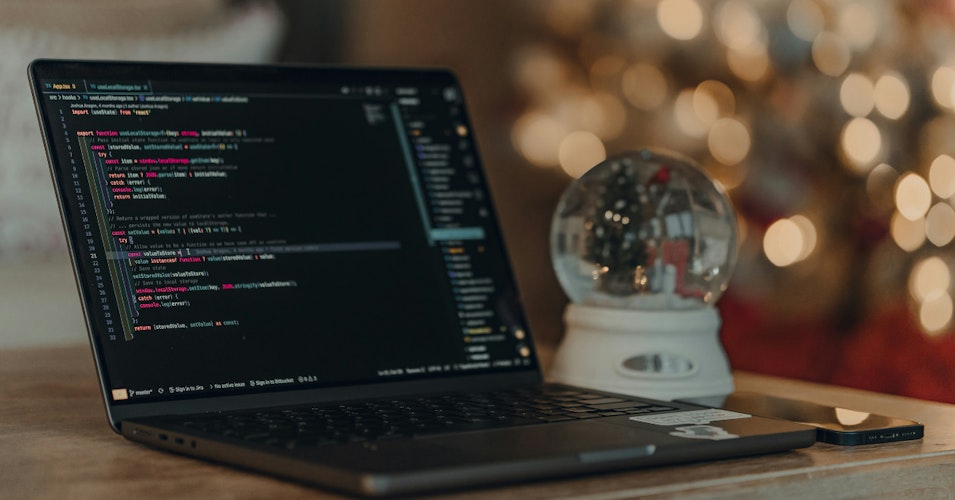 A laptop on a wooden table with a snow globe next to it and holiday lights in the background