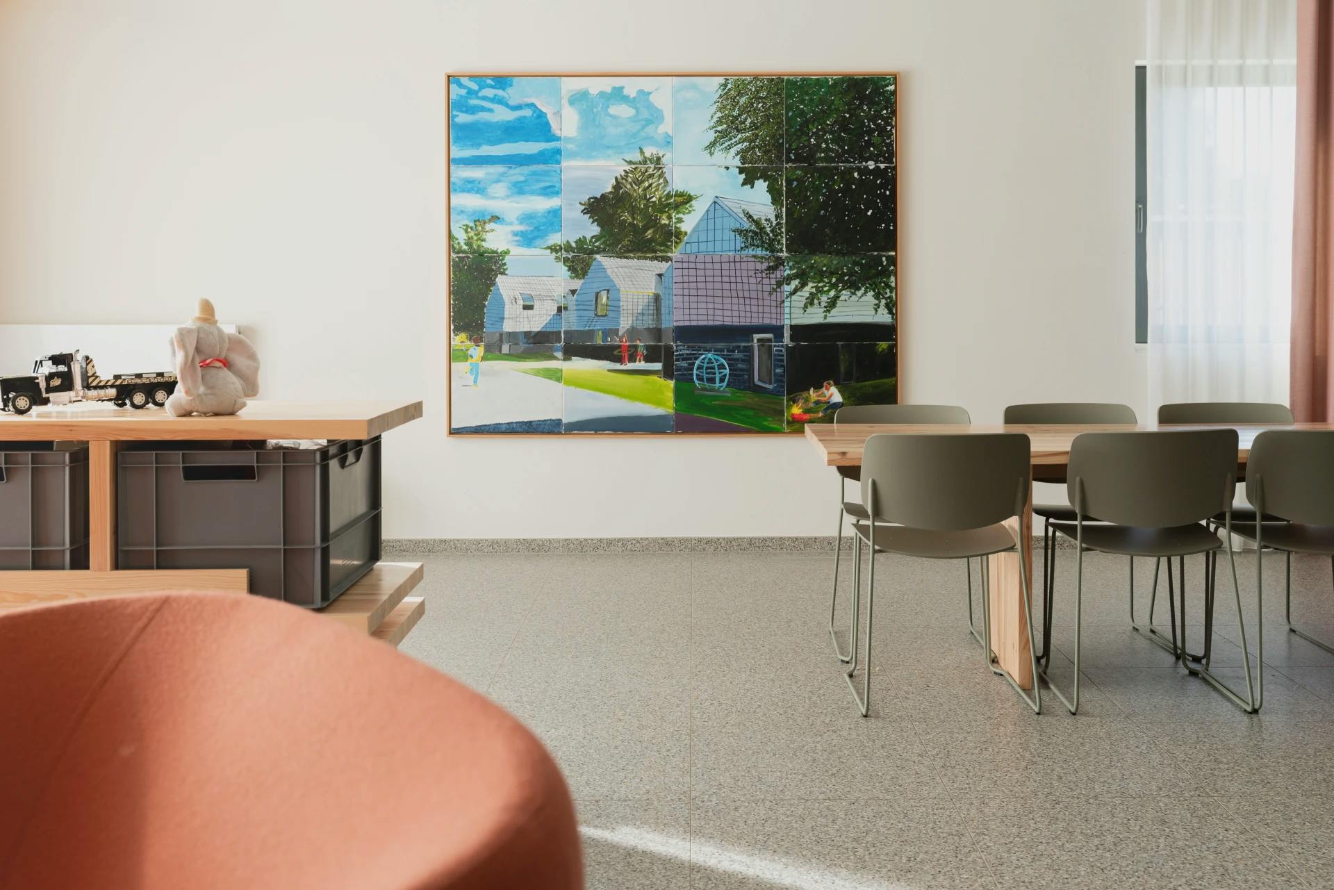 Kamer met schilderij van een huis en bomen, houten tafel, stoelen en speelgoed.