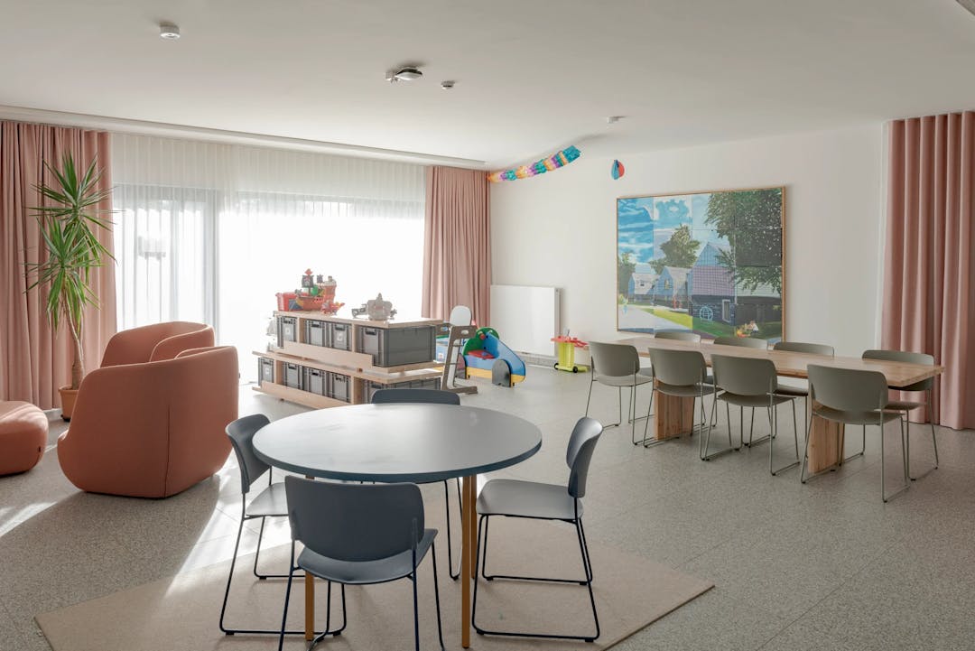 Ruime woonkamer met ronde en rechthoekige tafels, speelgoed en grote ramen met roze gordijnen.
