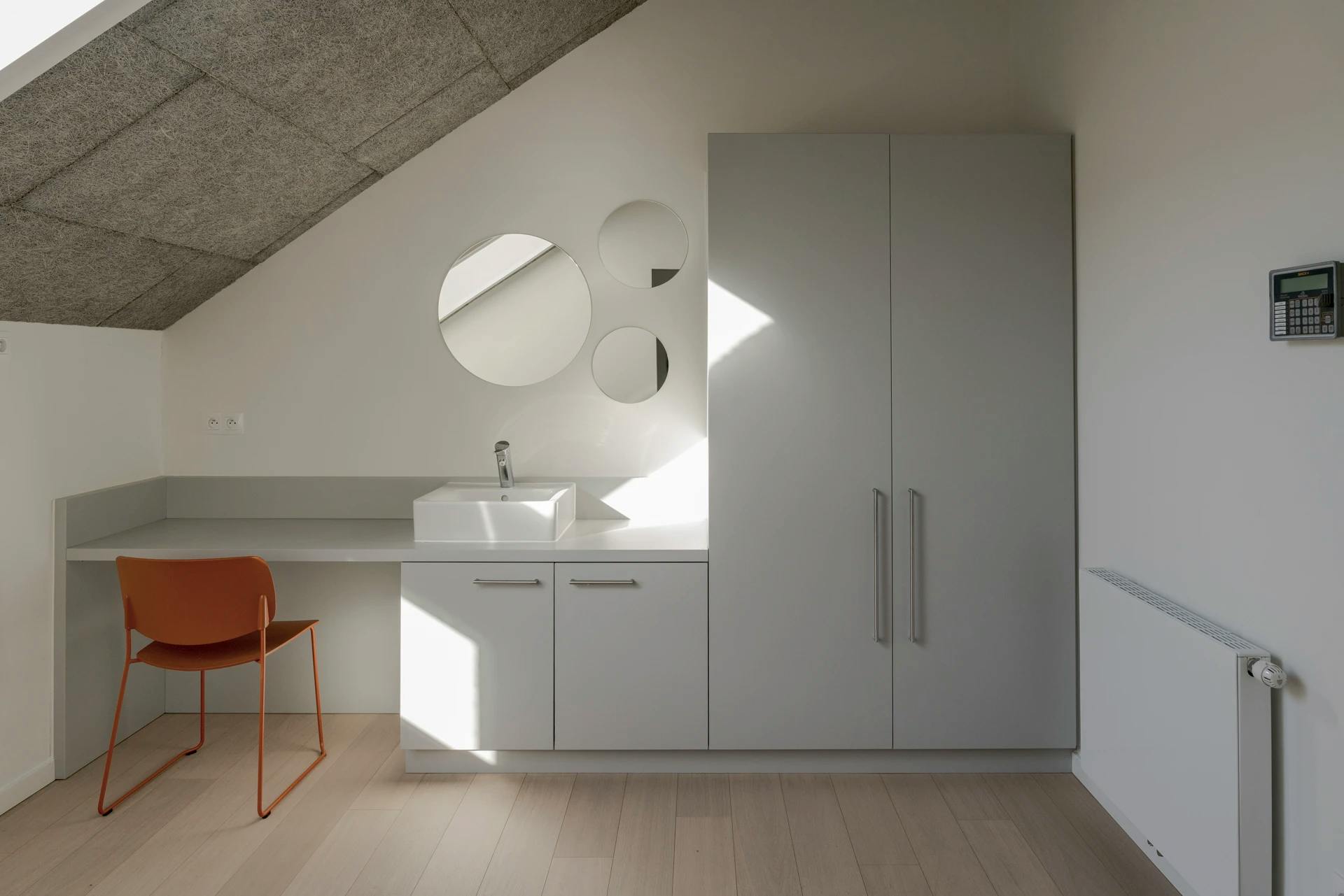 Minimalistische badkamer met witte wastafel, oranje stoel en ronde spiegels tegen een schuine wand.