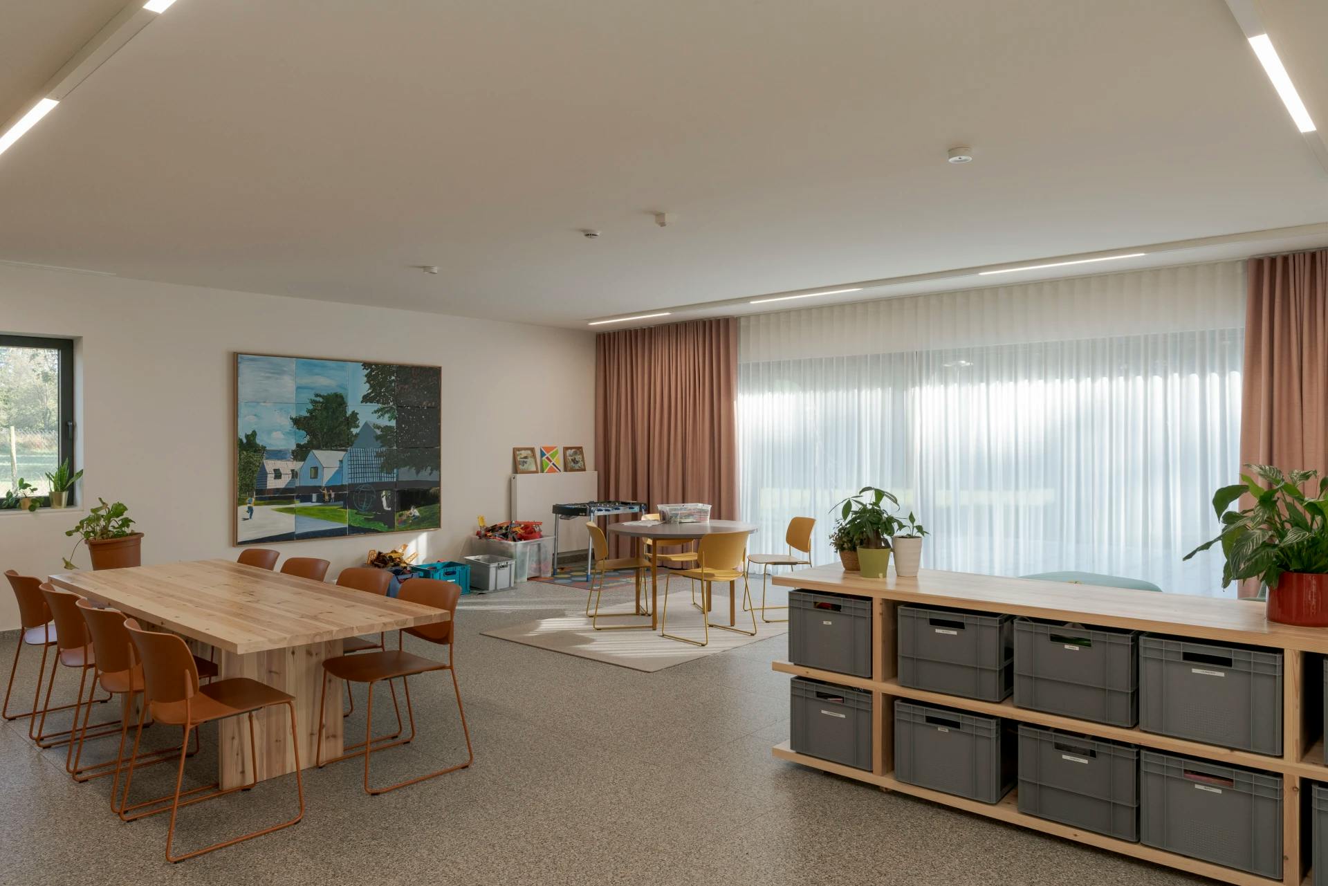 Multifunctionele ruimte met lange tafel, opbergbakken en speelgoed, omringd door veel licht en planten.