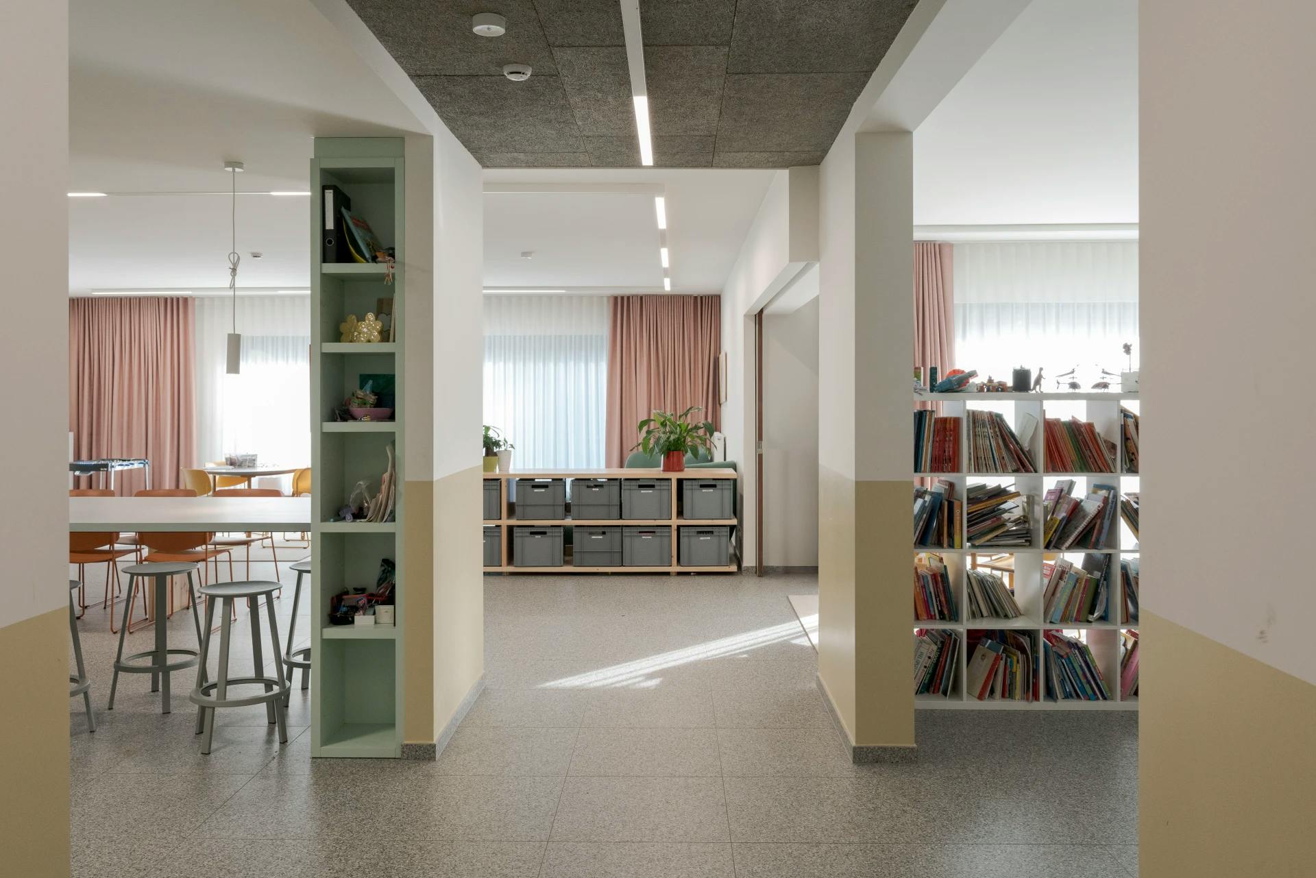 Gang met boekenrek en open zicht naar een ruimte met tafels en roze gordijnen.