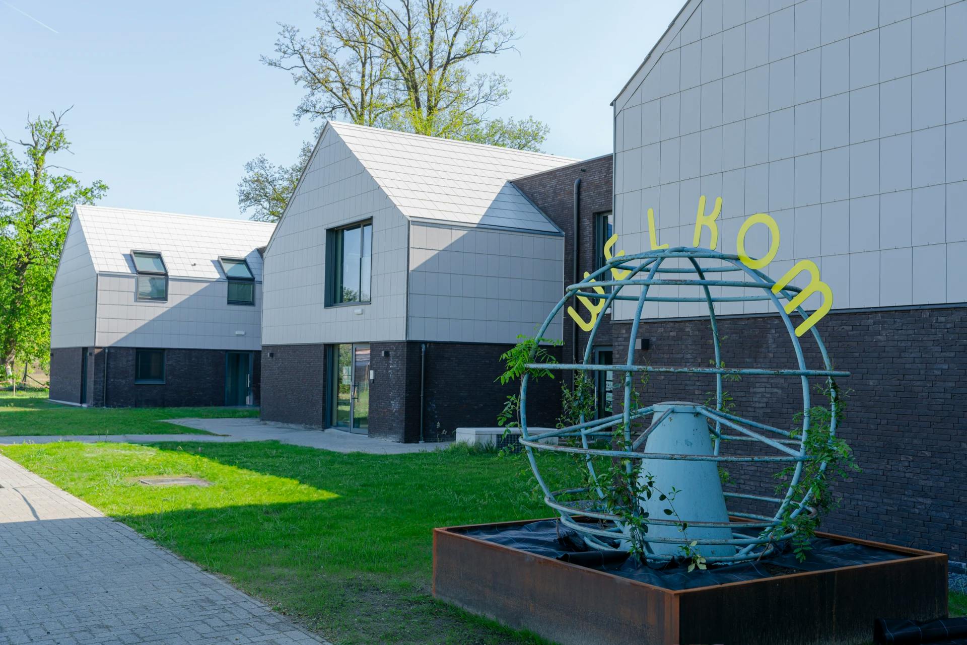 Welkom-installatie met gele letters bij moderne witte huizen, omlijst door gras en bomen.