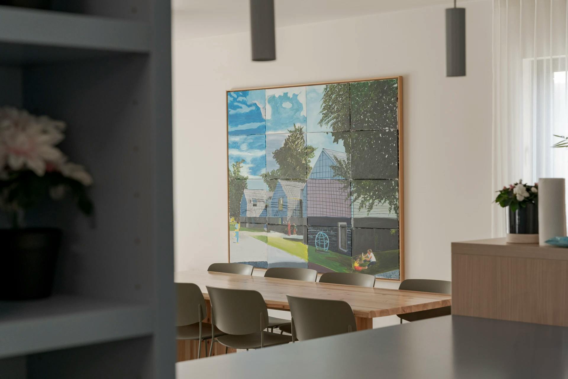 Schilderij van een modern huis met bomen, zicht vanaf een houten tafel en stoelen in een lichte ruimte.