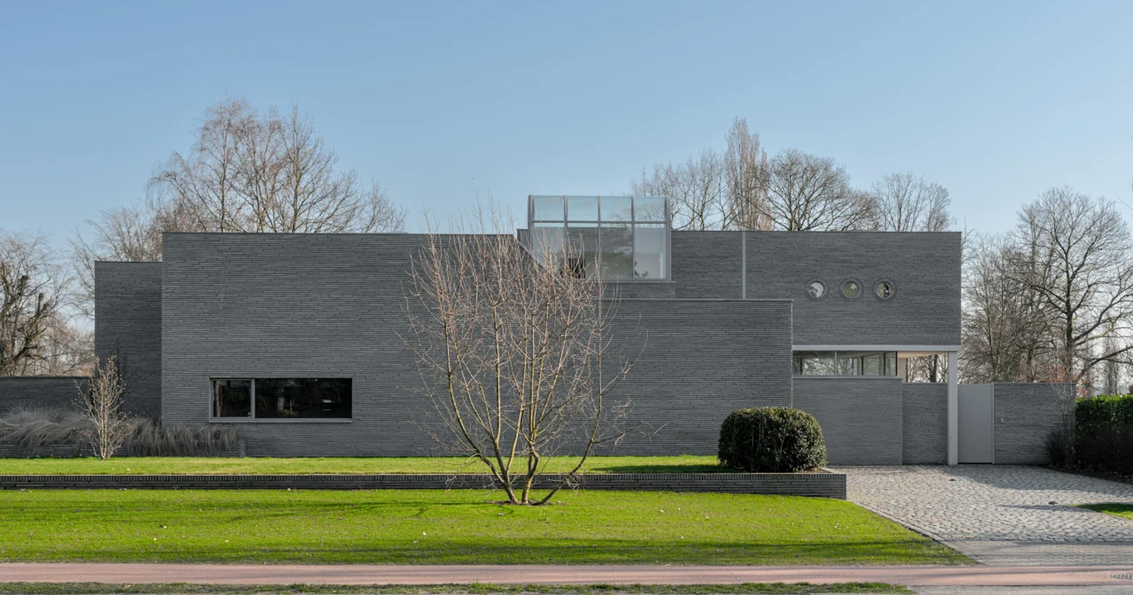Minimalistische woning met grijze bakstenen gevel, ronde ramen, een glazen uitbouw en een kale boom in de voortuin.