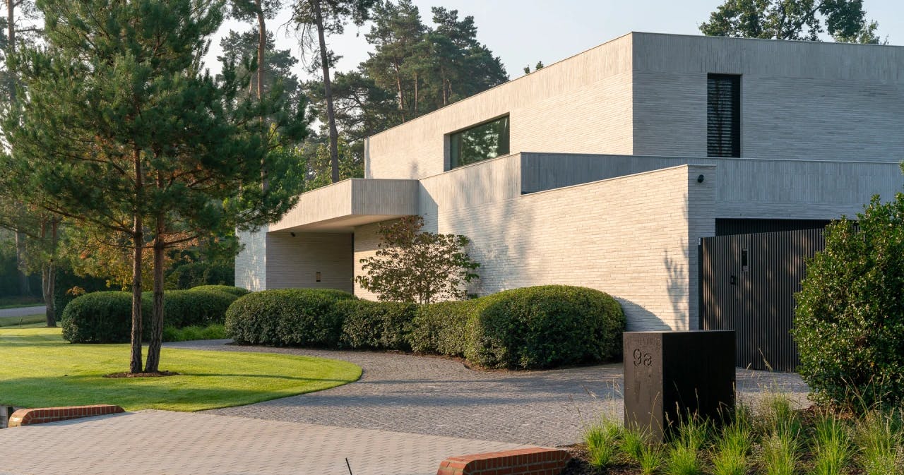 Modern huis met witte bakstenen gevels, grote heggen en een toegangsweg omgeven door bomen.