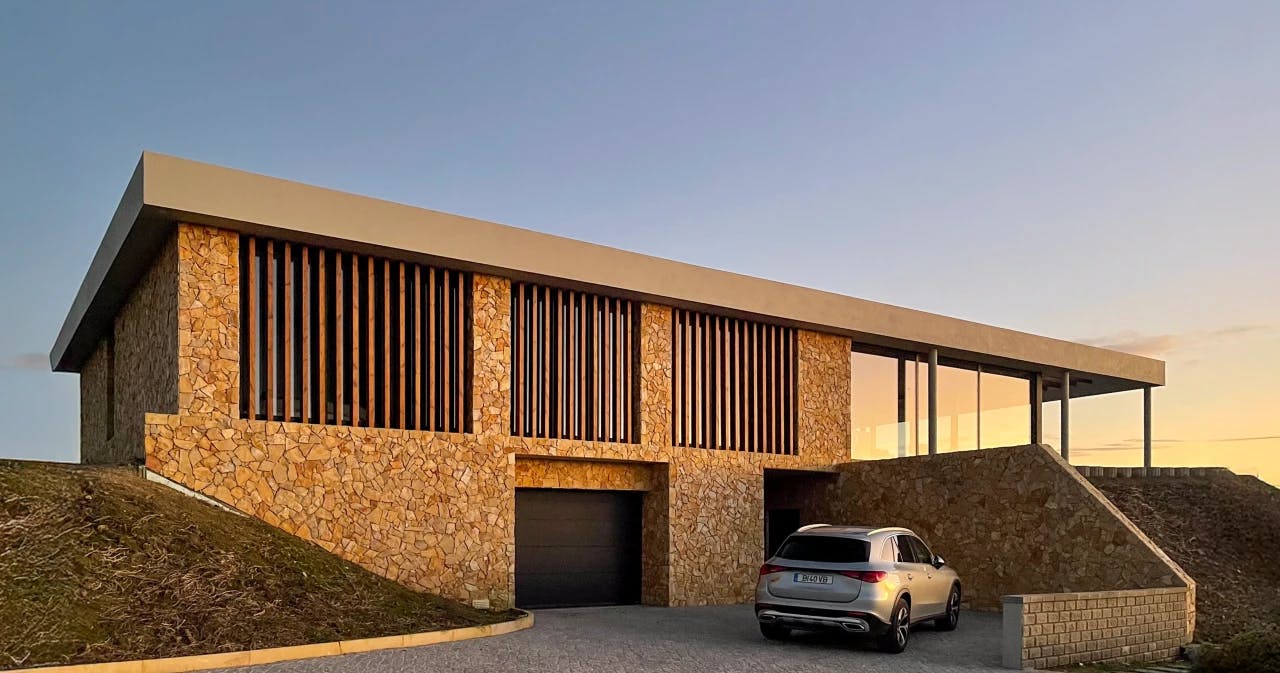 Voorgevel van een modern huis met natuurstenen muren, houten accenten en een oprit met een geparkeerde auto.