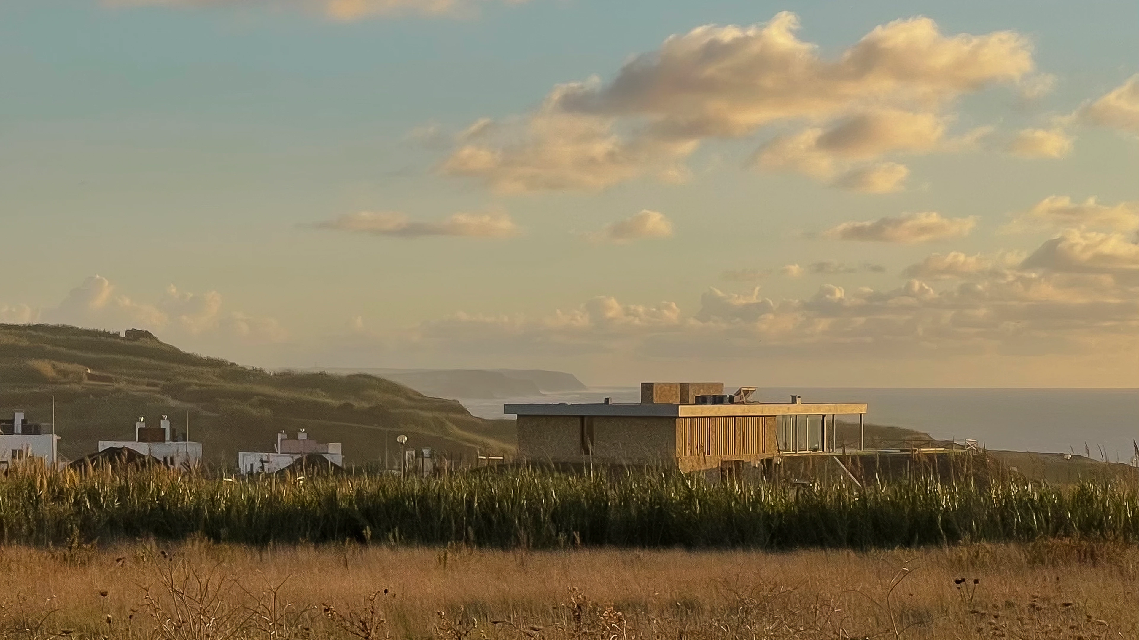 Modern huis met natuurstenen gevel, grote ramen en uitzicht op zee, omringd door heuvels en grasland bij zonsondergang.