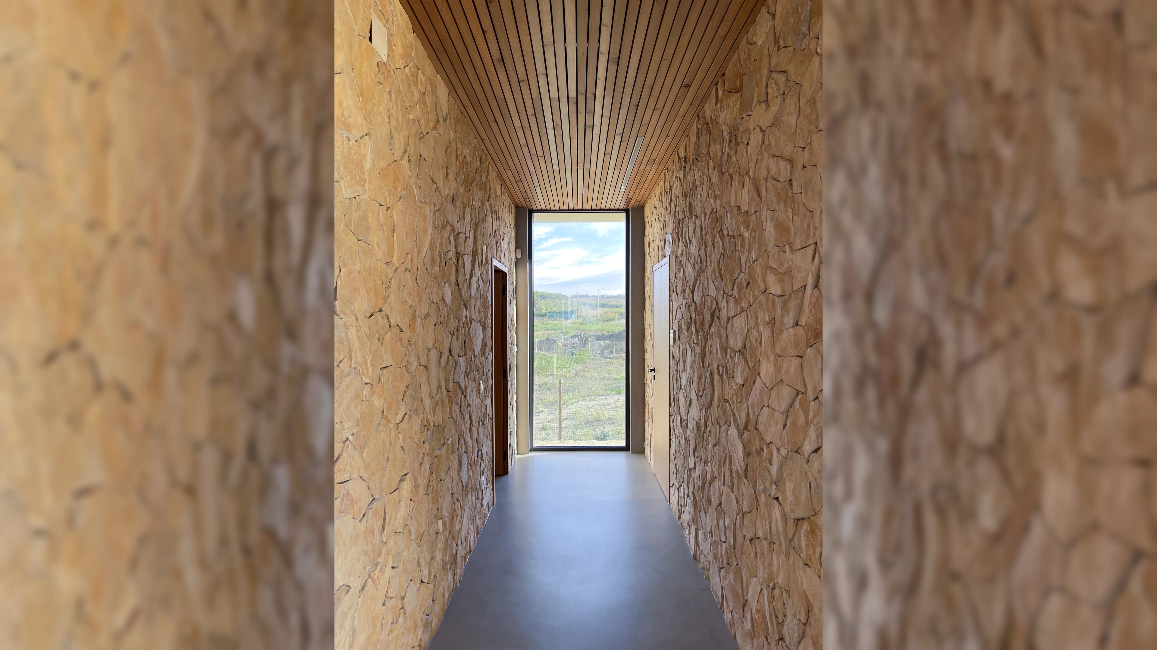 Smalle gang met natuurstenen muren, houten plafond en een groot raam dat uitzicht biedt op het landschap.