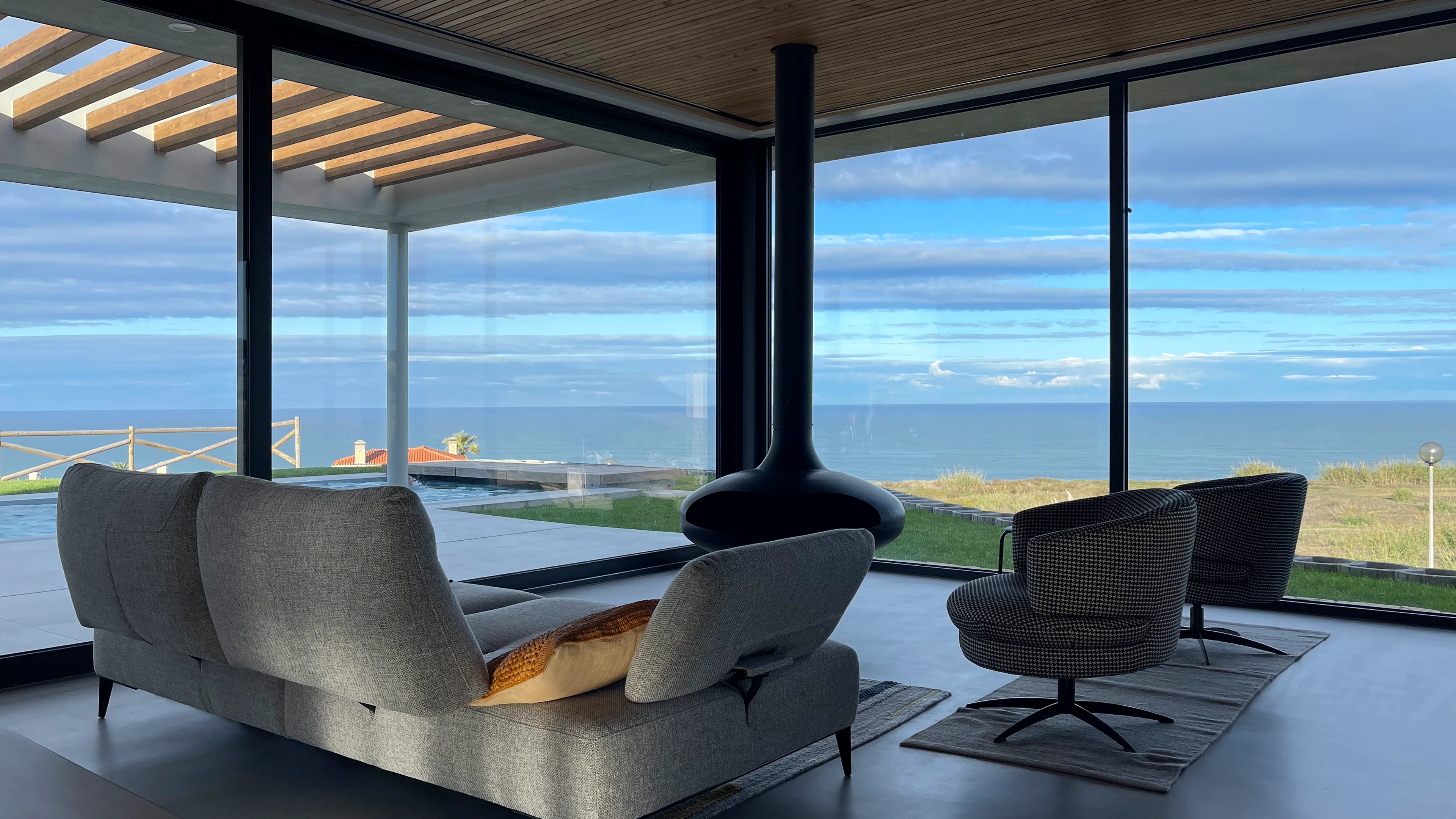 Woonkamer met glazen wanden, modern meubilair en uitzicht op zee, zwembad en een overdekt terras.
