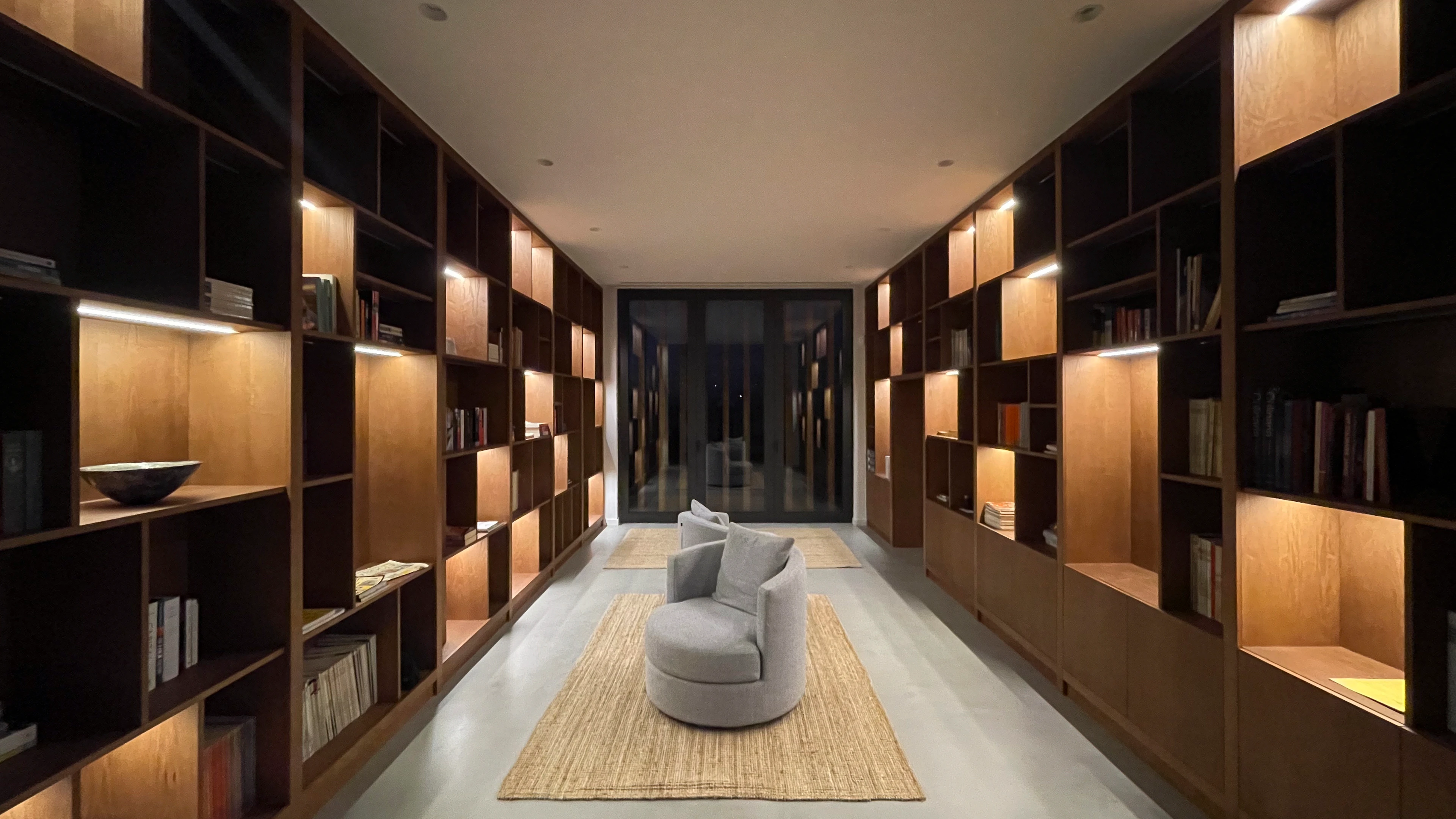 Sfeervol verlichte gang met houten kasten en een moderne fauteuil, leidend naar een glazen deur.