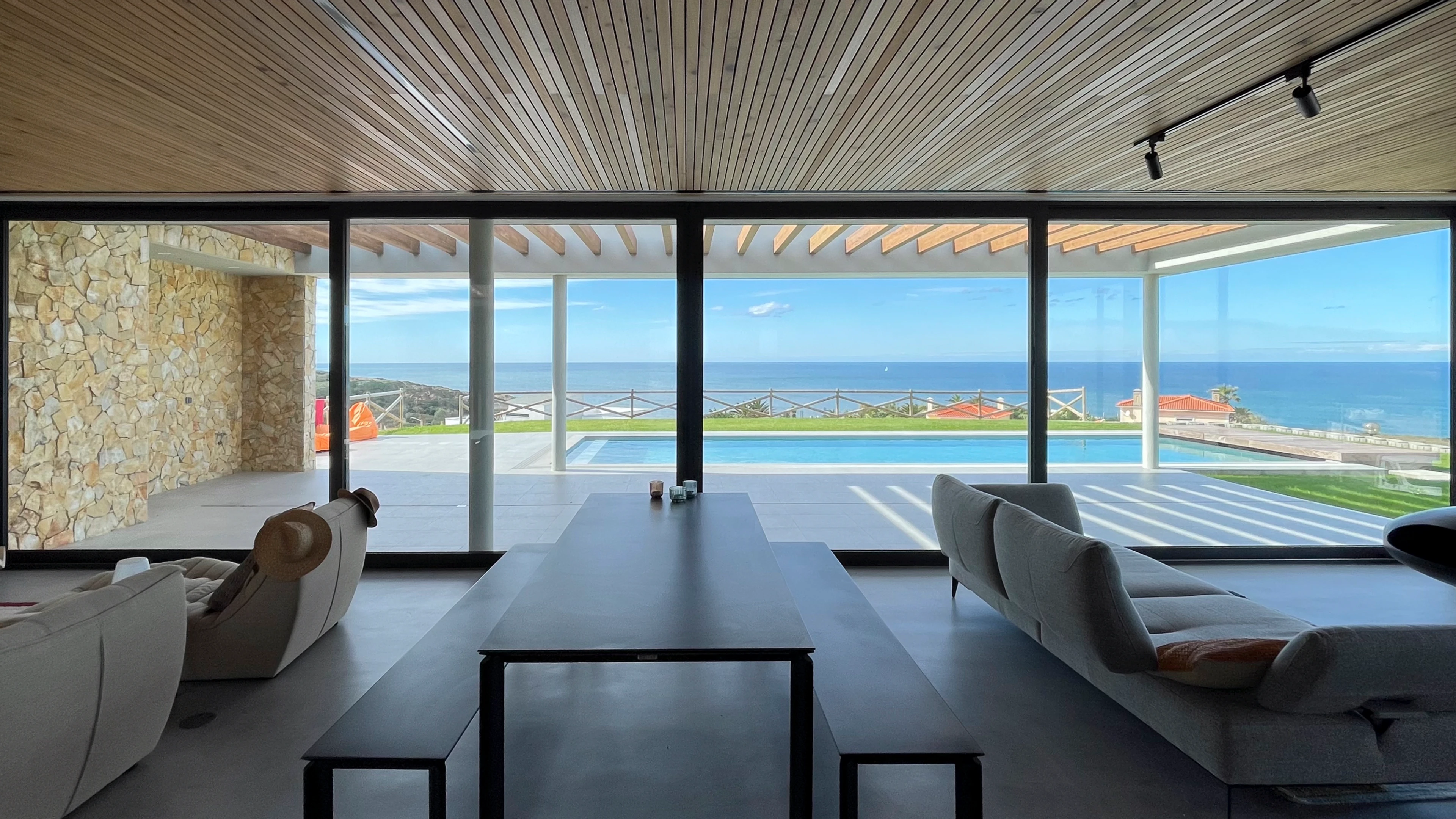 Binnenruimte met houten plafond, glazen wanden en een tafel, met uitzicht op een zwembad en de oceaan.