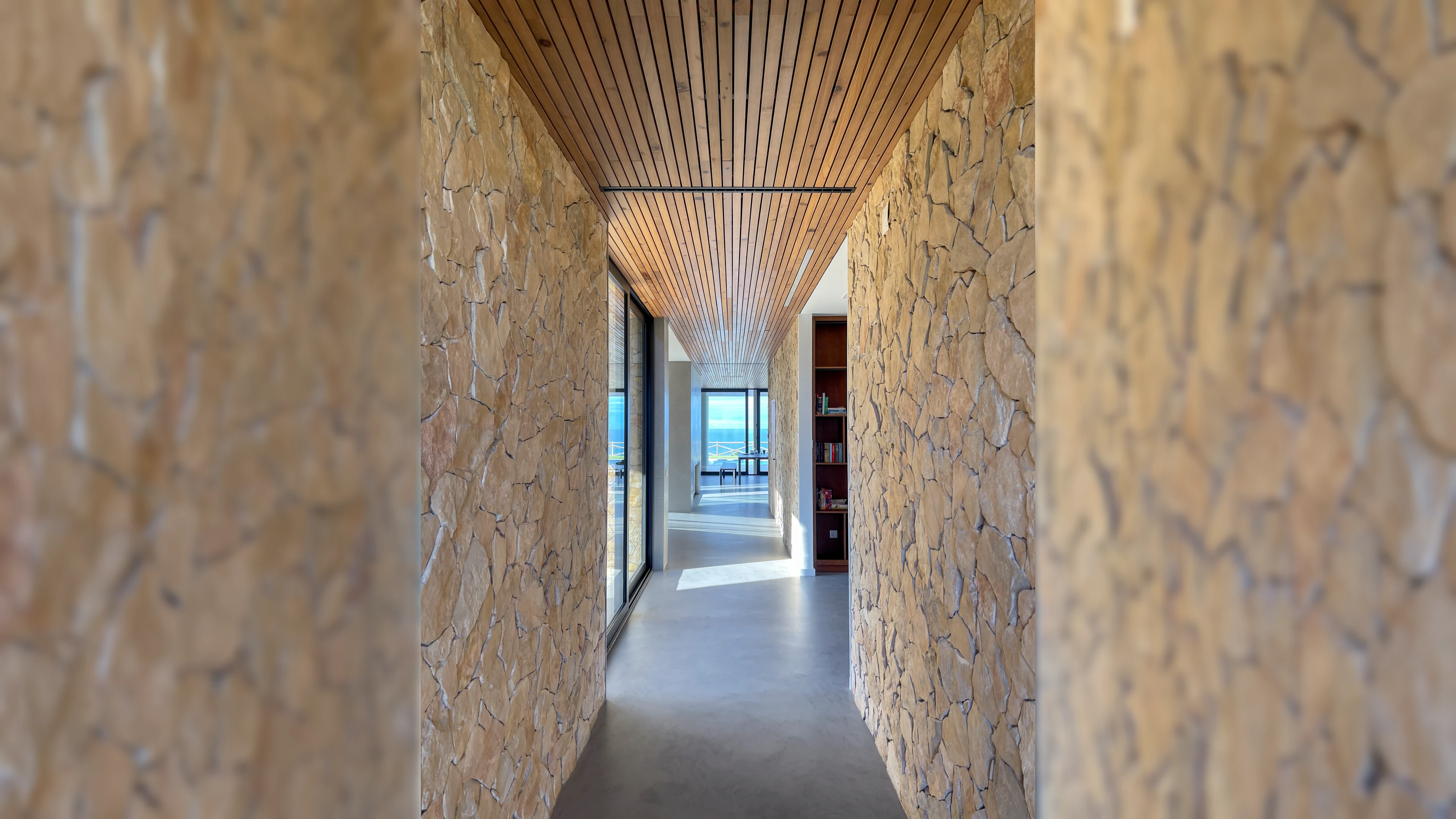 Gang met natuurstenen muren en houten plafond, eindigend bij een ruime woonruimte met uitzicht op zee.