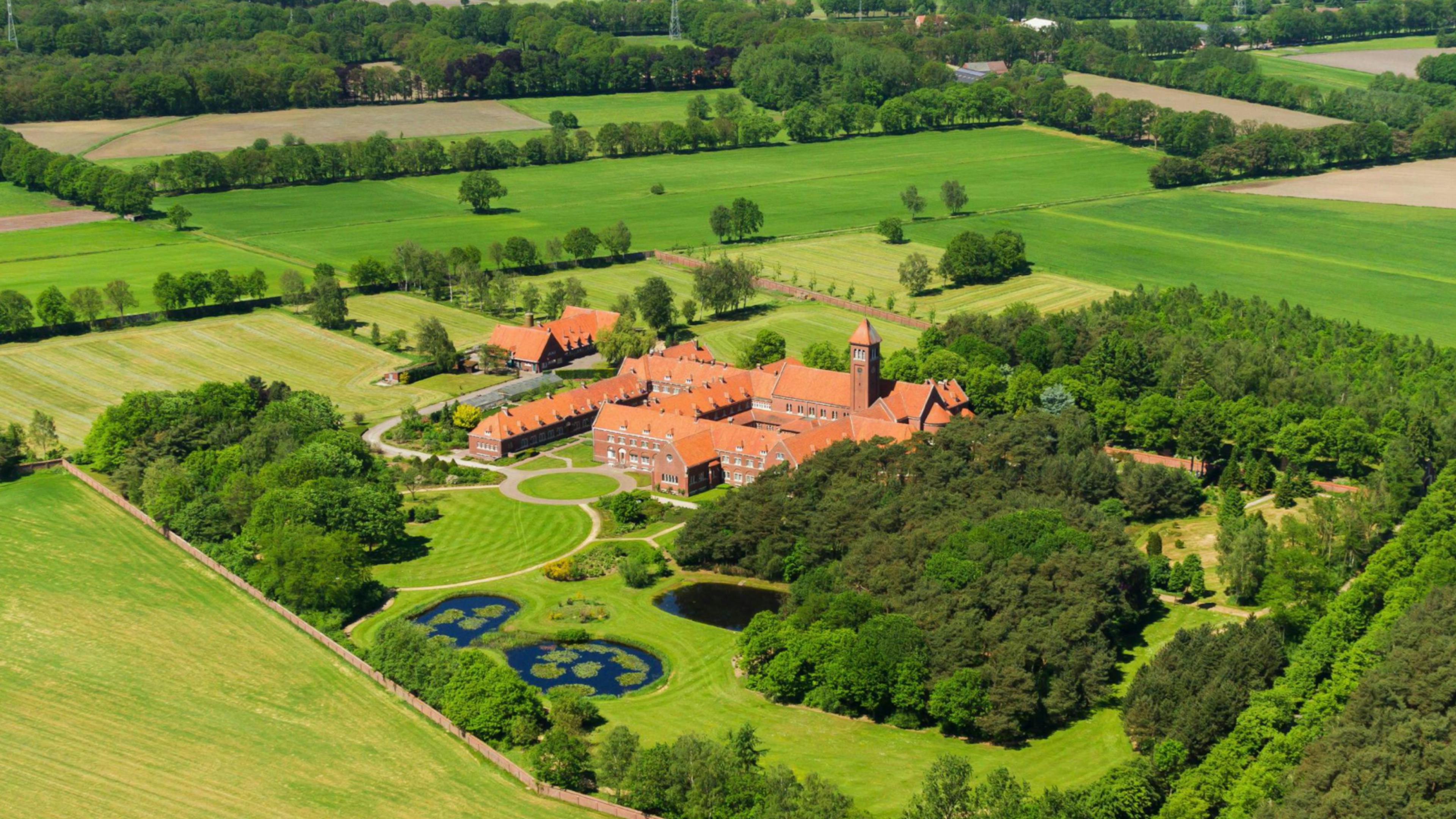 Luchtfoto van een abdijcomplex met rode daken, omringd door groene velden, bossen en vijvers.