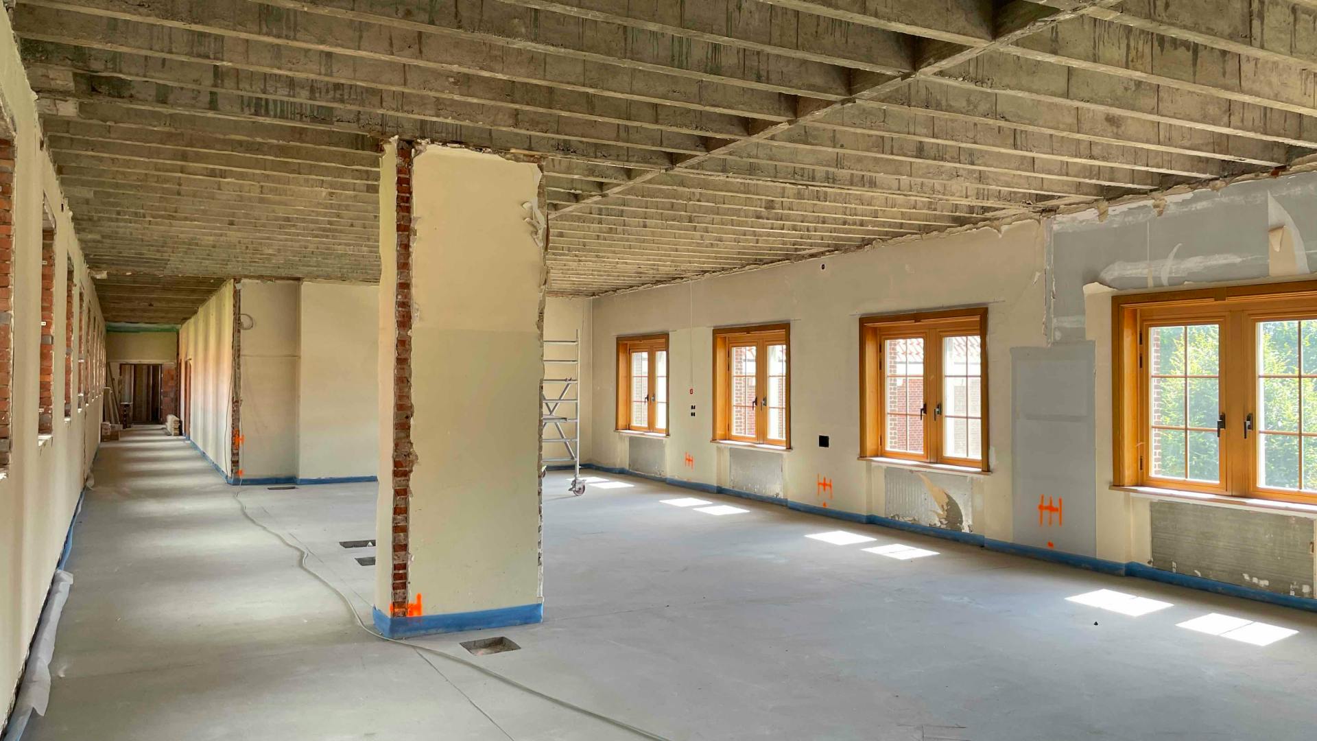 Grote ruimte in renovatie met ontmanteld plafond, onafgewerkte muren en houten raamkozijnen met uitzicht op een binnenplaats.