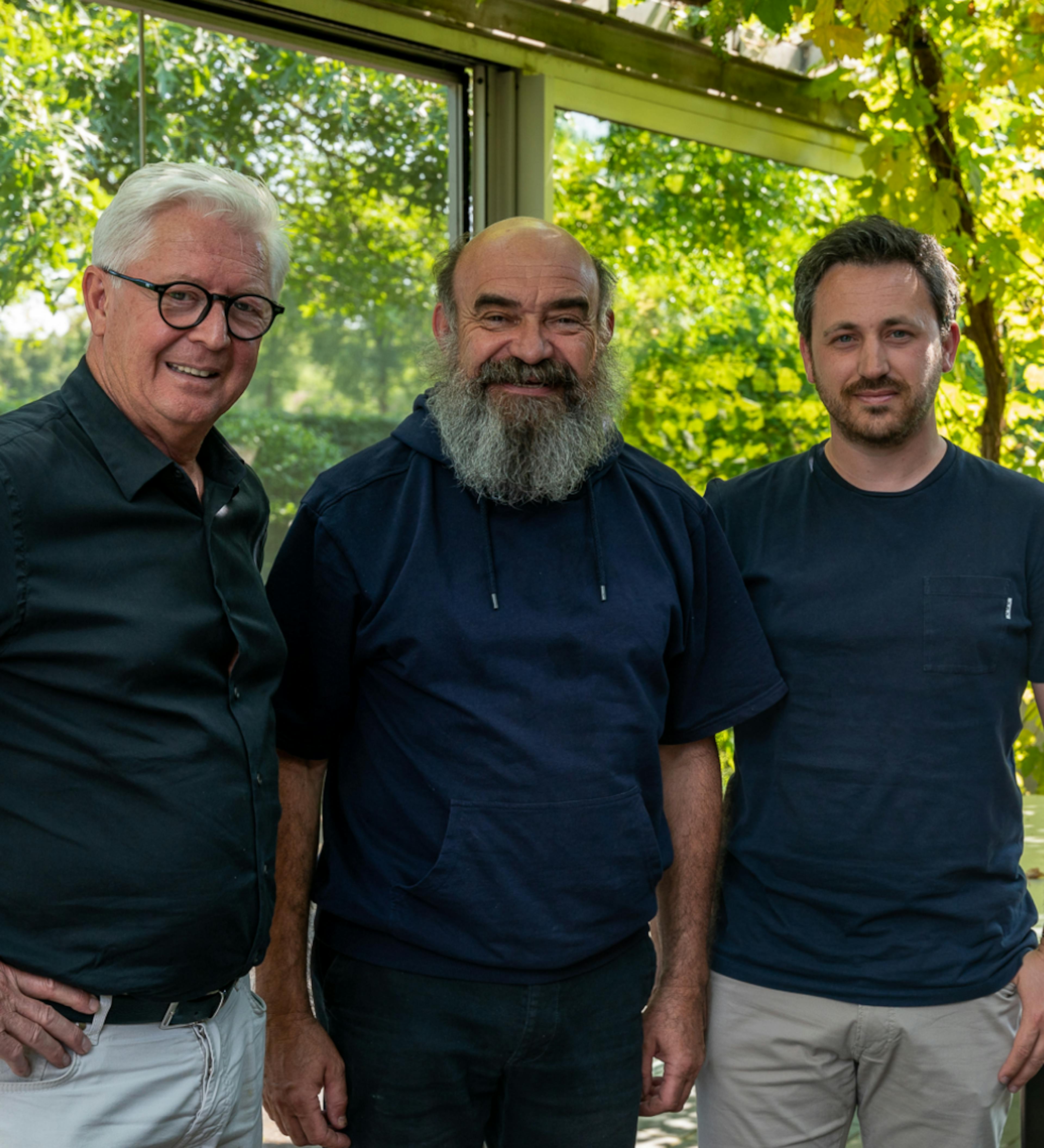 Drie mannen poseren lachend in een serre met groene planten op de achtergrond.