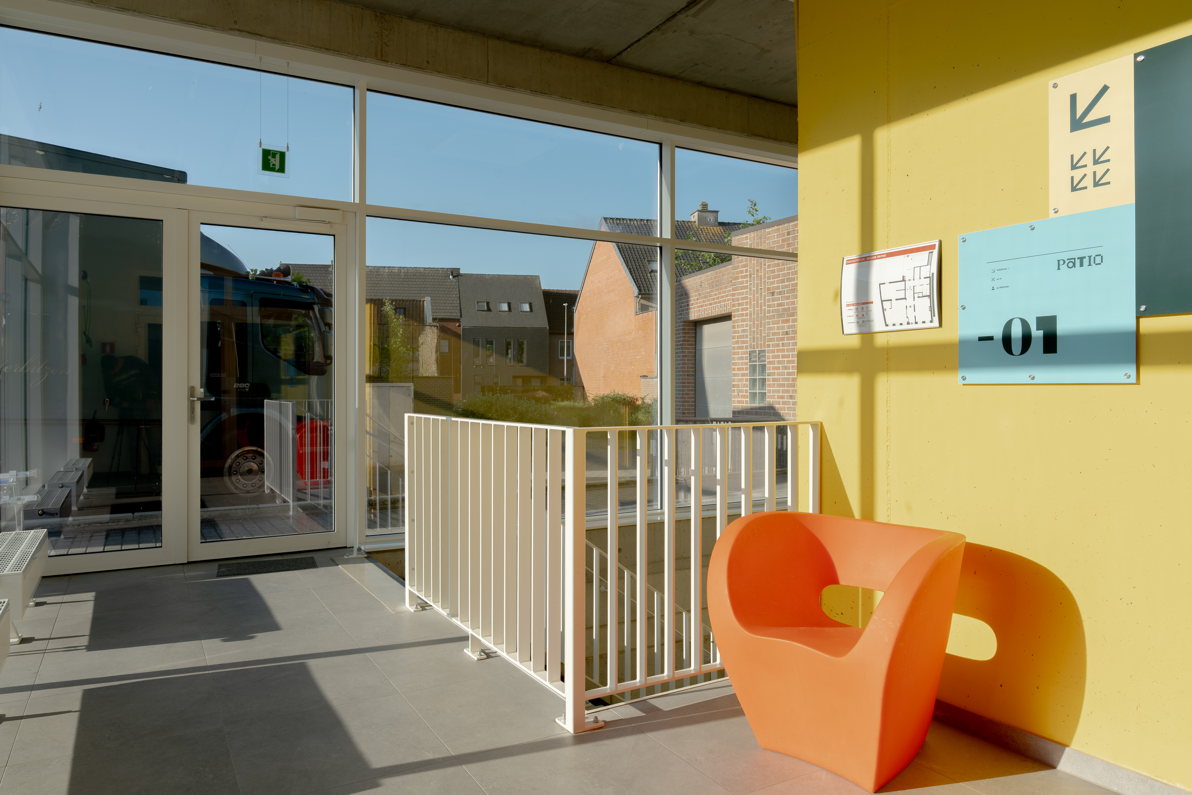 Binnenruimte met gele muur, oranje stoel en groot raam met uitzicht op straat.