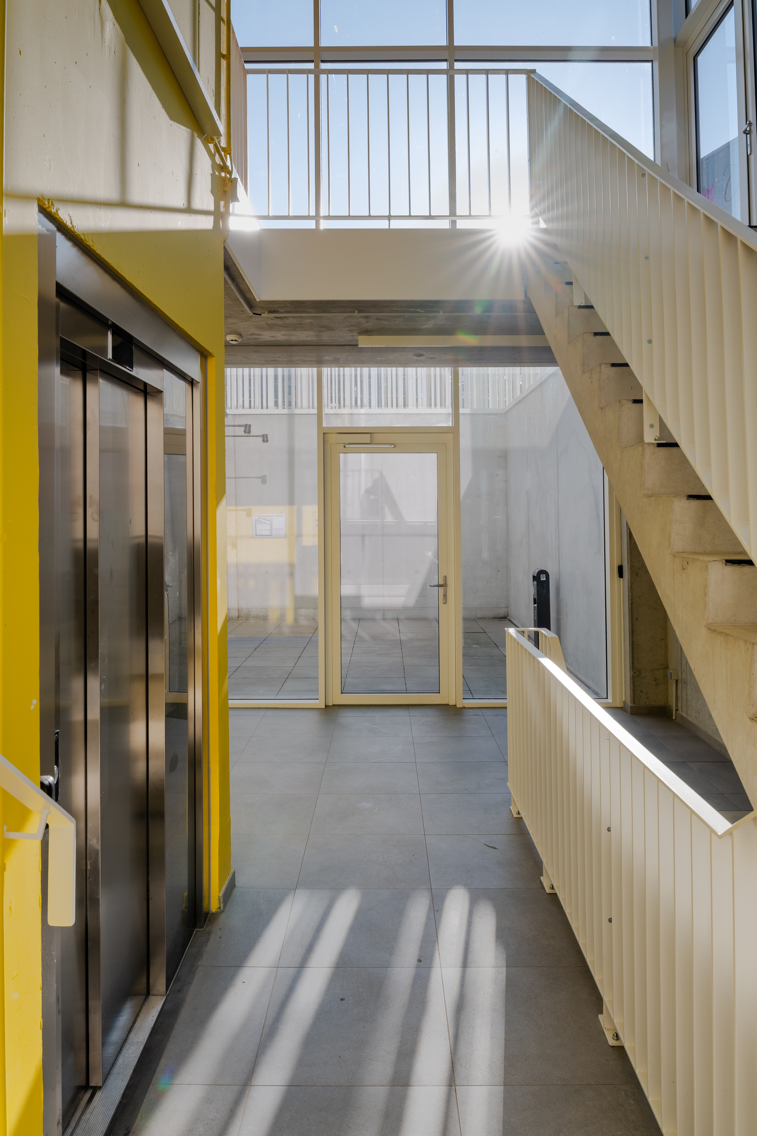 Hal met lift, trap en zonlicht dat door hoge ramen naar binnen valt.