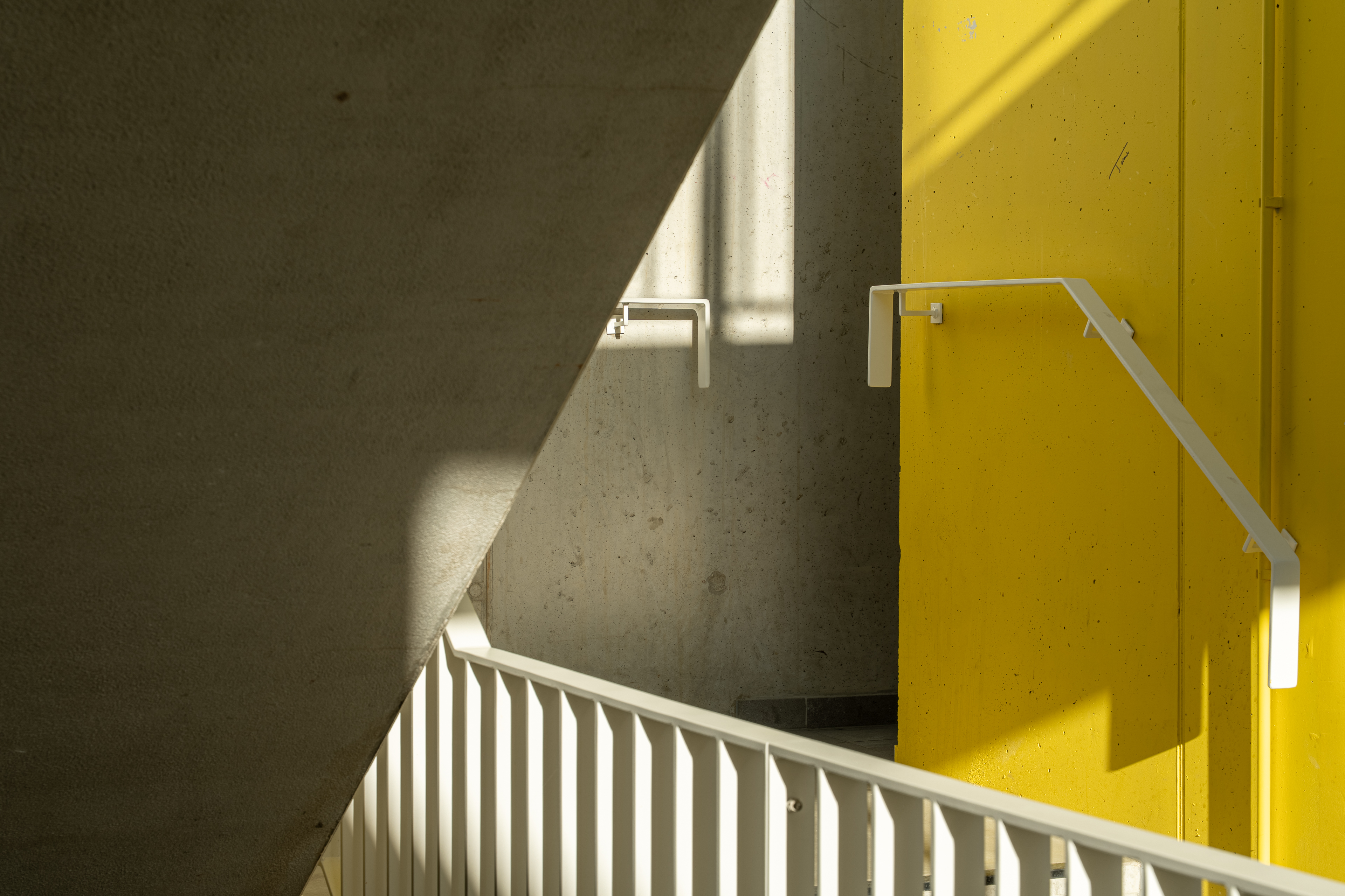 Trapdetail met betonnen muren, gele wand en witte leuning in zonlicht.