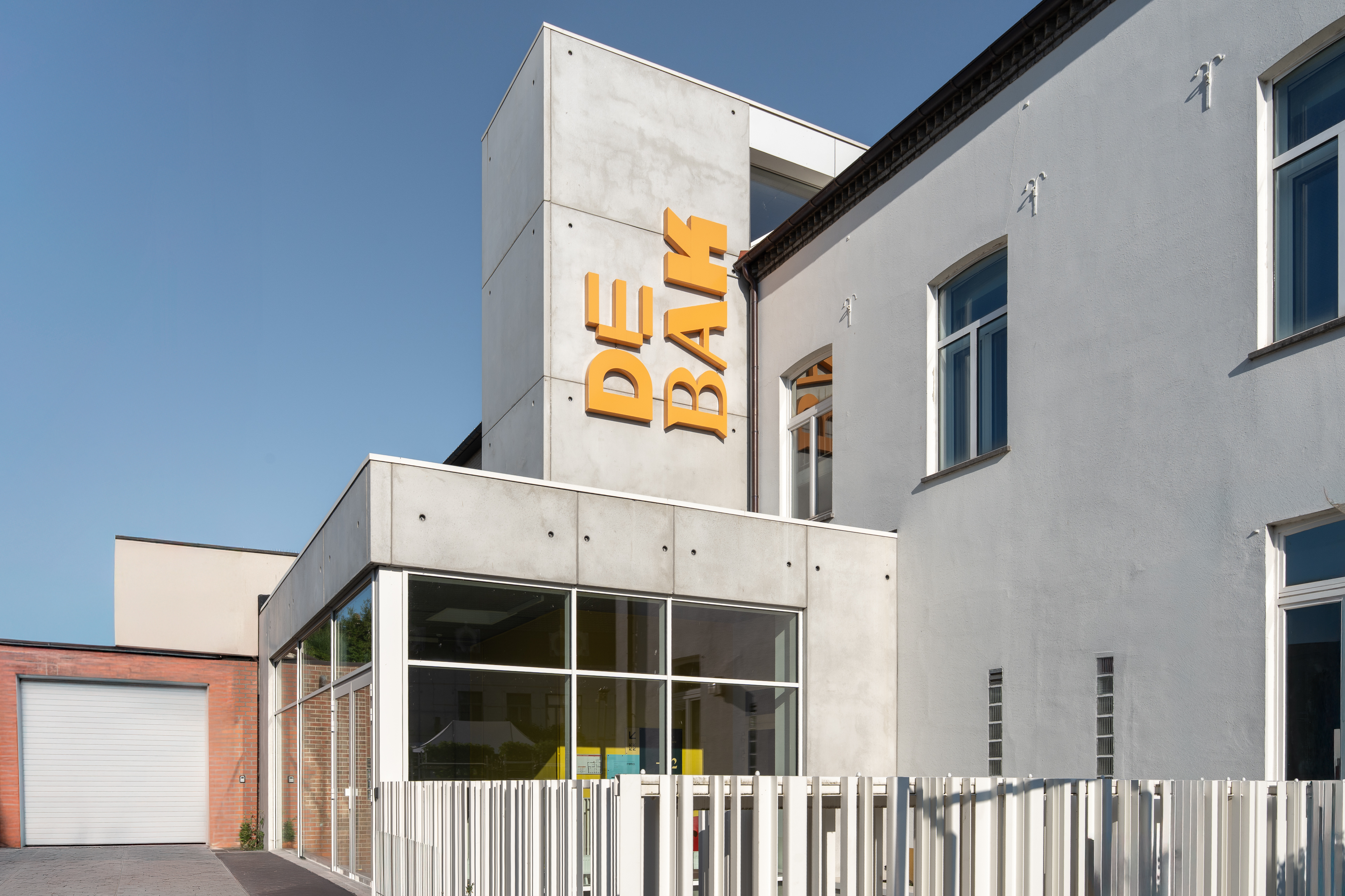 Voorkant van modern gebouw met betonnen gevel, grote ramen en gele letters DE BAK.