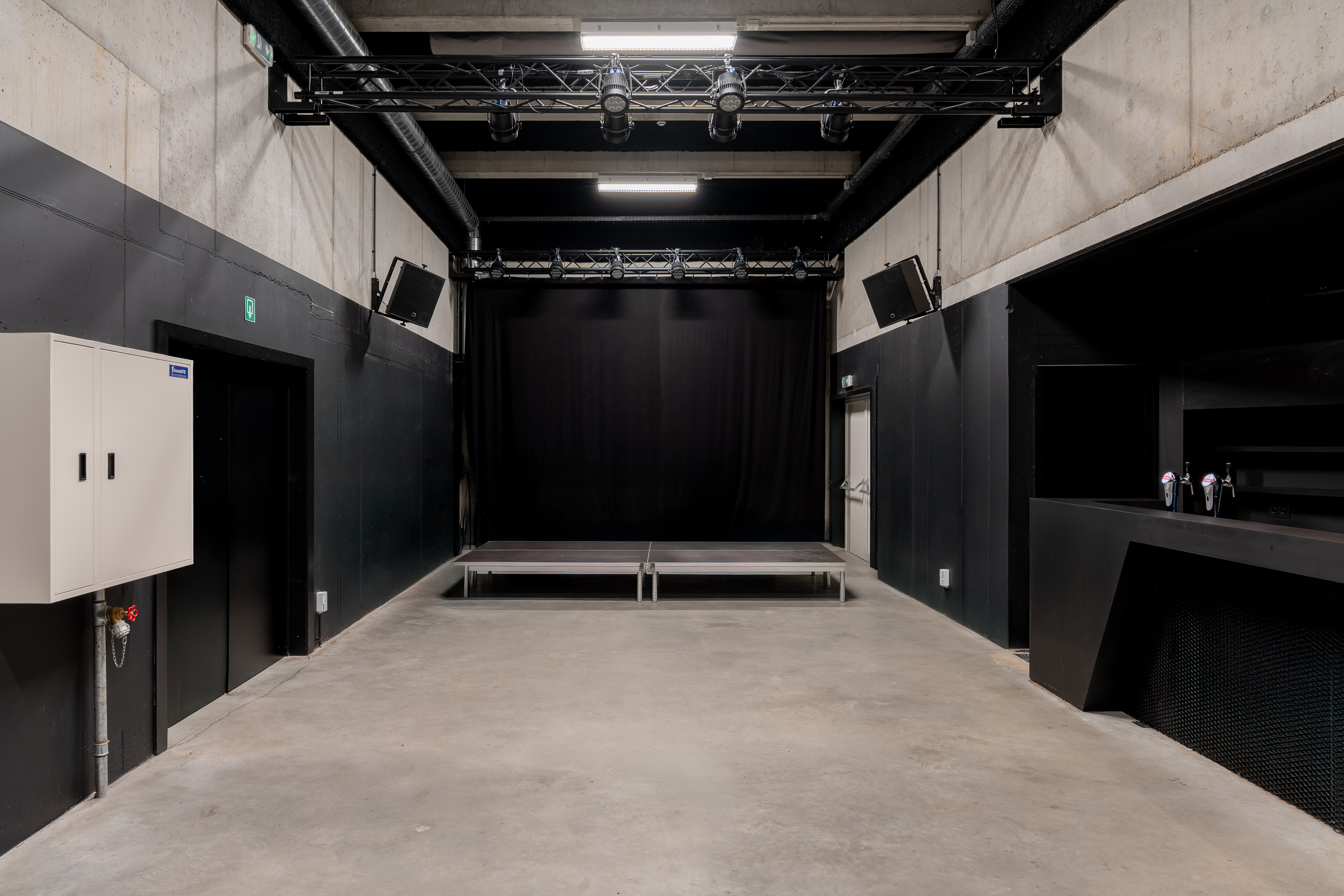 Zaal met klein podium, zwarte wanden en technische verlichting aan plafond.