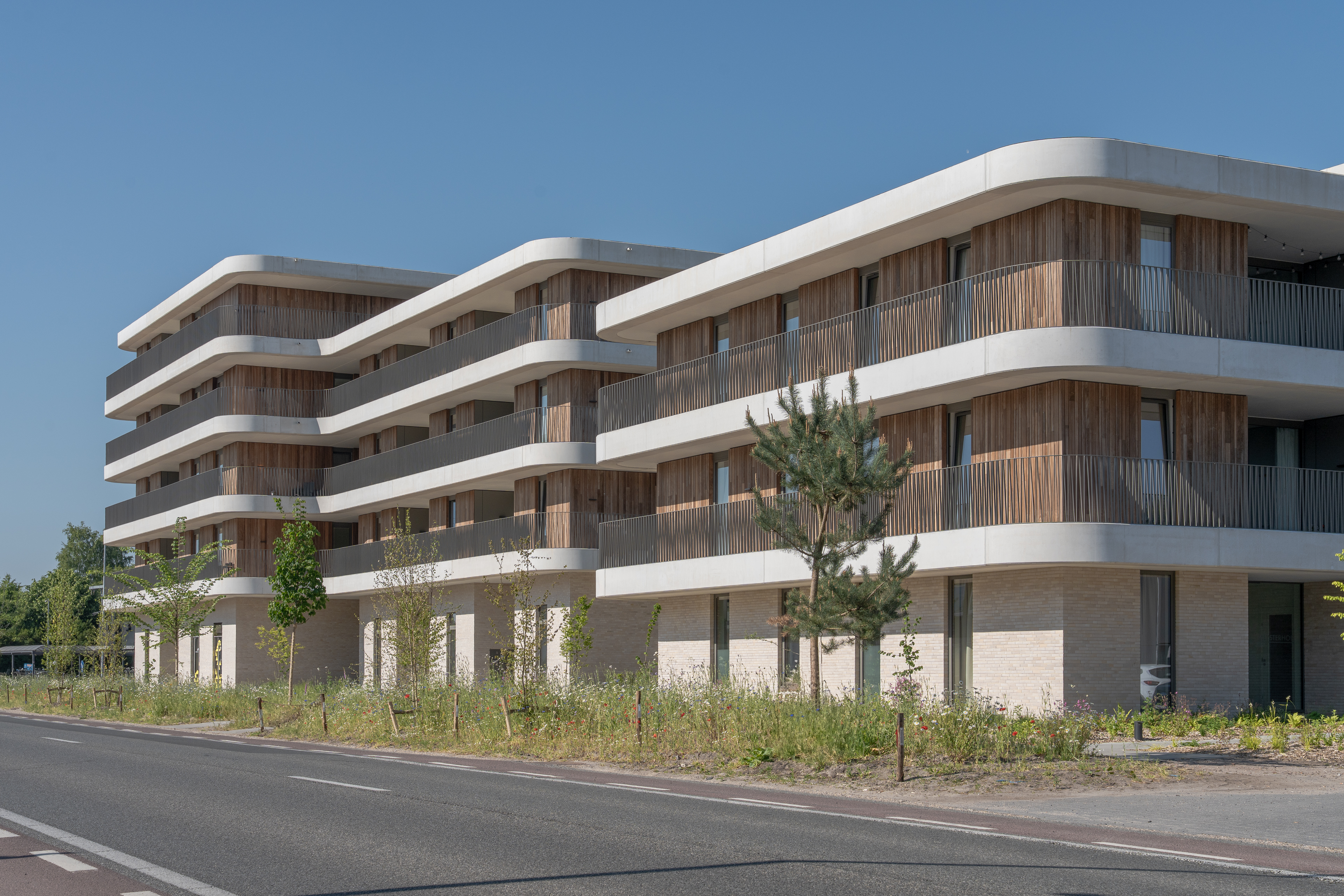 Modern appartementencomplex met ronde hoeken, houten accenten en brede balkons langs een weg.
